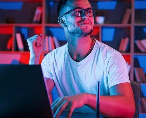 positive emotions. man in glasses and white shirt is sitting by the laptop in dark room with neon lighting